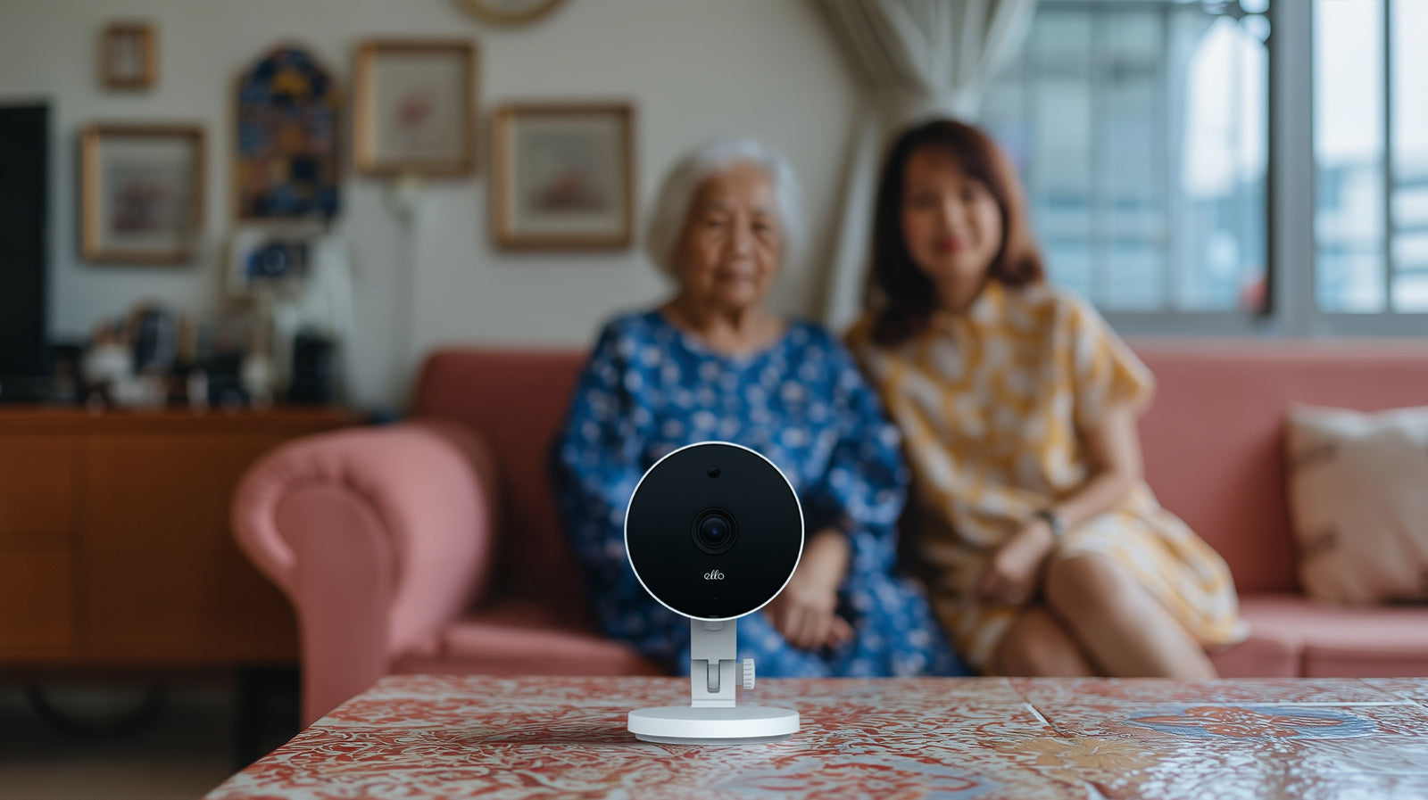 An ELLO camera on a table, with a mother and daughter sitting on a sofa behind it